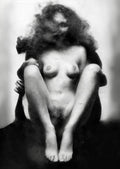 Black and white artistic photograph of a nude woman seated with her legs bent, surrounded by smoke or fog against a light studio background. Unmoored series.